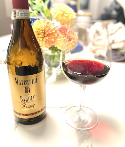A bottle of Marcini Barolo and a glass of red wine on a table with flowers in the background