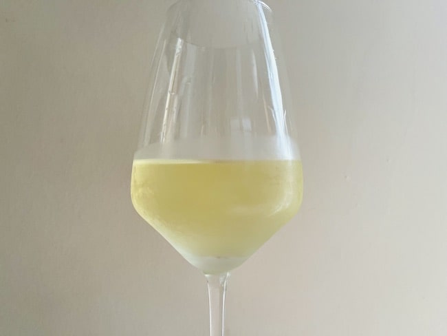 A close up of a wine glass half full of chilled white wine in front of a white wall