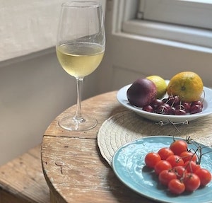 A glass of white wine on a wooden table with a plate of tomatoes and a bowl of cherries and lemons