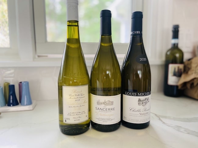 Three bottles of white French wines that go with oysters Sancerre, and Chablis on a white marble kitchen countertop with a window in the background