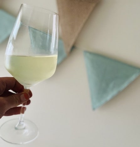 A hand holds a glass of white wine on an angle in front of a wall with pastel bunting